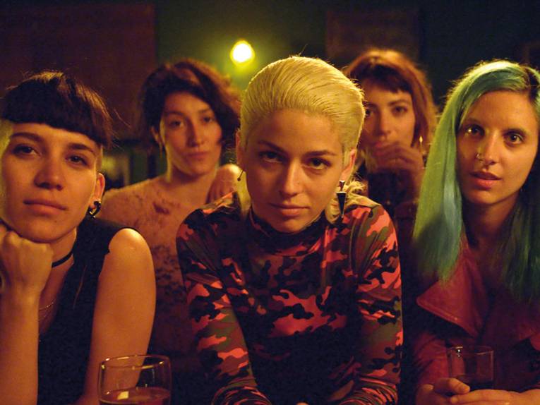 Auf dem in dunklen gelb-orang-Tönen gehaltenen Foto sieht man vorne in der Mitte Ema. Sie hat gelb-blonde nach hinten gegelte Haare, trägt ein langärmeliges Shirt mit Tarnmuster und guckt direkt in die Kamera. Neben und hinter ihr stehen weitere vier Frauen, die in die Kamera gucken.